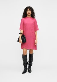 Ροζ διαφανές φόρεμα με μανίκια μήκους ως τον αγκώνα, συνδυασμένο με μια μαύρη τσάντα με υφή και μαύρες μπότες μέχρι το γόνατο, τοποθετημένα πάνω σε λευκό φόντο.