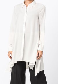 Blusa bianca a maniche lunghe con chiusura a bottoni, orlo fluido e tessuto liscio e leggero. Abbinata a pantaloni neri.