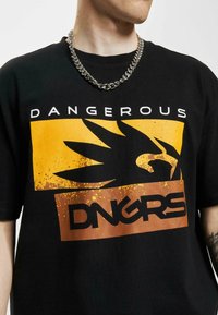 Sort bomulds T-shirt med et markant gult og brunt grafisk design, tekst der læser "DANGEROUS" og "DNGRS," samt et tekstureret kæderegnhalskæde.