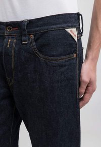 Jeans de mezclilla oscura con un corte clásico, con costuras naranjas, una etiqueta roja en el bolsillo trasero y una textura suave.