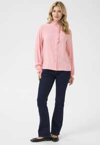 Light pink long-sleeve blouse with frill detail and button front, paired with dark blue flared jeans and black flat shoes.