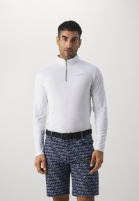Hvit langermet zip-up overdel med ståkrage, kombinert med marineblå shorts med et gjentakende hvitt logo-mønster. Teksturen fremstår som jevn.