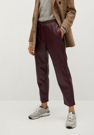 Persoon draagt een bruin ruitjesblazer, een grijs kabelgebreid trui, bordeauxrode leren broek met trekkoord en grijze sneakers, staand op een witte vloer.