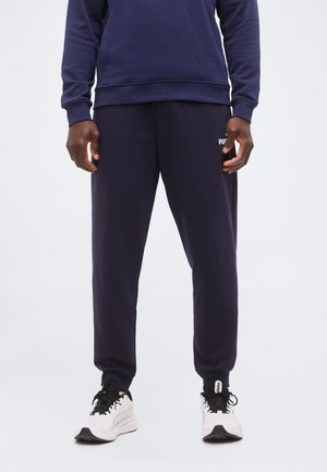 Homme portant un pantalon de survêtement bleu marine Puma et un sweat assorti avec des baskets blanches Puma, debout devant un fond clair uni.