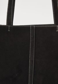 Geantă tote din suede negru, cu cusături proeminente, formă rectangulară și două mânere rezistente. Textură netedă cu un design minimal.