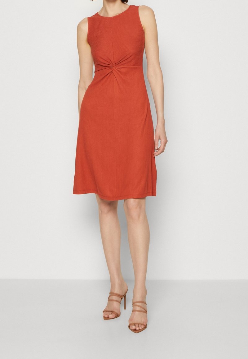 Robe sans manches de couleur rouille avec un corsage ajusté, détail en torsion à la taille et jupe évasée. Texture lisse ; design longueur genoux.