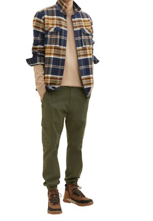 Persona che indossa un maglione beige sotto una camicia a quadri blu e arancione, pantaloni cargo verde oliva e scarponi da trekking marroni, in piedi con le mani nelle tasche.