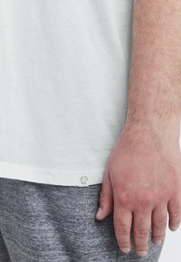 Hvid bomulds t-shirt med et rundt mærke ved kanten, parret med grå teksturerede joggers. Stoffet ser blødt og letvægtigt ud.