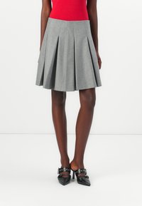 Hame gris plissé en tissu texturé, avec une taille haute et une forme évasée, associé à des chaussures noires pointues avec des accents de bouclerie.