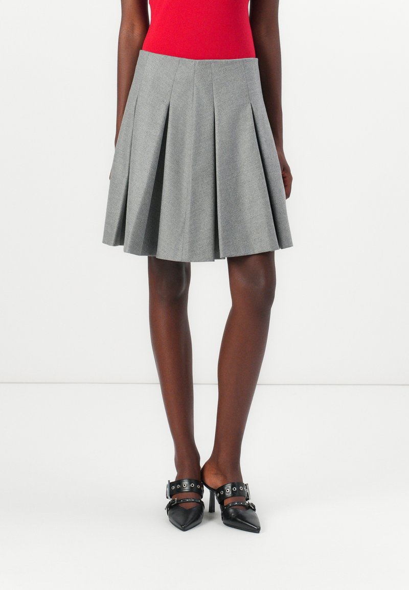 Hame gris plissé en tissu texturé, avec une taille haute et une forme évasée, associé à des chaussures noires pointues avec des accents de bouclerie.