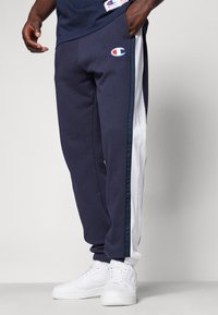 Pantalones de chándal azul marino con rayas laterales blancas, puños elásticos y un parche de logo en la parte delantera izquierda. Fabricados con un tejido suave y texturizado.