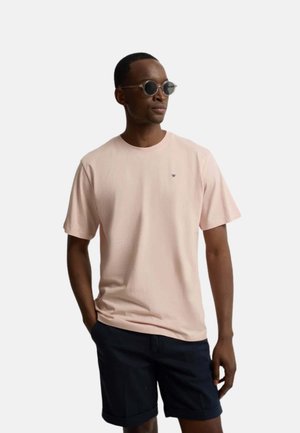 Hombre con gafas de sol redondas, camiseta rosa claro de manga corta y pantalones cortos azul marino oscuro, de pie con una mano en el bolsillo.