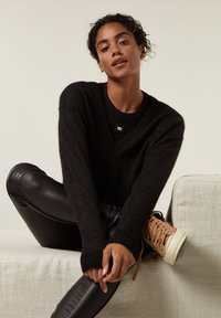 Zwarte, chunky gebreide trui met een V-hals, gecombineerd met zwarte neplederen leggings. Gekenmerkt door tan hoge sneakers met bruine veters.