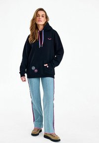 Naine, kes kannab musta kapuutsiga pusa, millel on roosad ja sinised nöörid, helesiniseid pükse roosade küljeribadega ning pruune matkasaapaid, seisab ühe käega taskus.