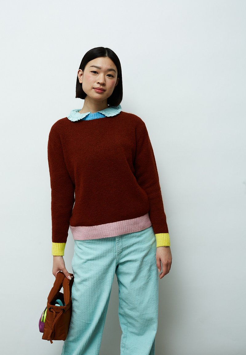 Pull rouille avec un ourlet rose et des poignets jaunes, porté sur une chemise bleue à col. Pantalon en velours côtelé bleu clair, tenant un sac marron.