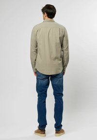 Camisa de manga larga beige con un corte relajado, detalles de botones en los puños, combinada con jeans de mezclilla azules y zapatos marrones, vista desde la parte posterior.