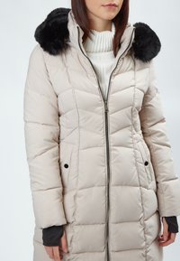 Cappotto invernale imbottito di un beige chiaro con cappuccio in pelliccia sintetica nera. Presenta una zip frontale, tasche laterali e un maglione bianco a costine sotto.