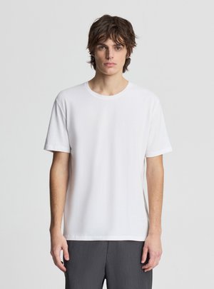 Giovane uomo con capelli castani di media lunghezza che indossa una maglietta bianca semplice a maniche corte e pantaloni grigio scuro su sfondo chiaro.