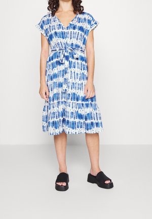 Femme portant une robe bleu et blanc à motifs, longueur genou, manches courtes avec une ceinture nouée à la taille, associée à des sandales compensées noires sans lacets.