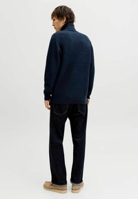 Dunkelblauer Rippenstrickpullover mit hohem Kragen, getragen über dunklen Jeans. Modell steht mit dem Rücken zur Kamera und zeigt die strukturierte Stoffqualität und die lockere Passform.