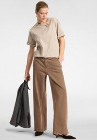 T-shirt beige a maniche corte con una spilla decorativa, abbinata a pantaloni larghi marrone chiaro, che tiene in mano un blazer grigio e sandali alti marrone scuro.