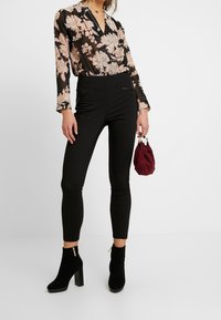 Czarna kwiecista bluzka z długimi rękawami, ozdobiona beżowymi kwiatami, w zestawieniu z czarnymi dopasowanymi spodniami i czarnymi botkami, trzymająca bordową torebkę.