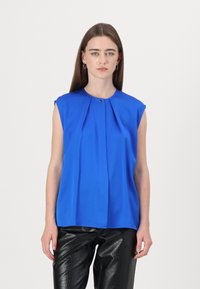 Blusa azzurra senza maniche con design plissettato, scollatura rotonda e piccolo accento dorato al colletto. Indossata con pantaloni neri testurizzati.