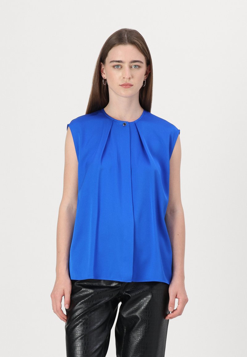 Blusa azzurra senza maniche con design plissettato, scollatura rotonda e piccolo accento dorato al colletto. Indossata con pantaloni neri testurizzati.