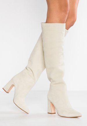 Vrouw die kniehoge, beige suède laarzen met blokhakken draagt, geposeerd met één been licht omhoog tegen een witte achtergrond.