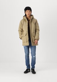 Khaki parka jakna s kapuljačom, dva prednja džepa i detaljima s čipkama. Uparena s plavim poderanim trapericama i crnim tenisicama.