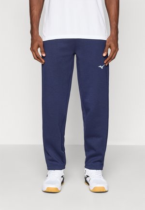 Homme portant un pantalon de survêtement bleu marine avec un petit logo sur la cuisse gauche et des chaussures de sport blanches avec des semelles en gomme, debout sur un sol neutre.