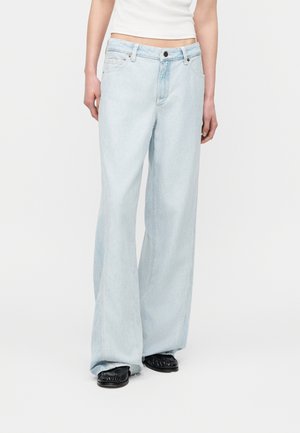 Άτομο που φοράει ανοιχτό μπλε τζιν με φαρδιά πόδια και μαύρα επίπεδα παπούτσια, στέκεται μπροστά από ένα απλό λευκό φόντο.