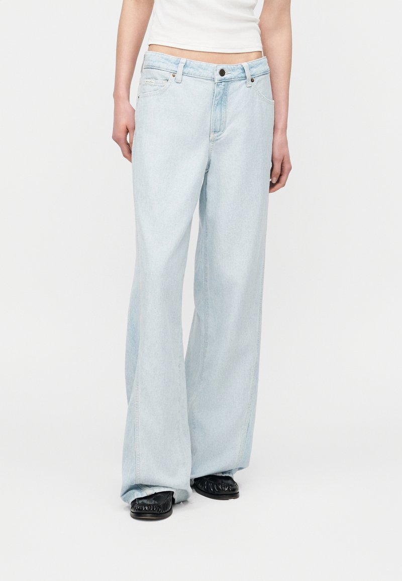 Άτομο που φοράει ανοιχτό μπλε τζιν με φαρδιά πόδια και μαύρα επίπεδα παπούτσια, στέκεται μπροστά από ένα απλό λευκό φόντο.