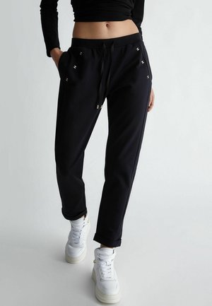 Pantalones deportivos negros con corte cónico, cintura con cordón y detalles metálicos decorativos en los bolsillos. Combinados con zapatillas blancas.
