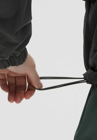 Crna jakna s elastičnom manžetom, koja ima vezicu u struku. Ruka drži dva crna užeta, naglašavajući funkcionalni dizajn.