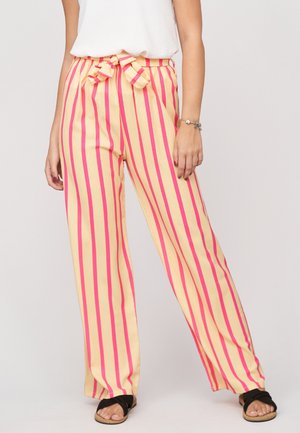 Mujer con pantalones de cintura alta y pernera ancha con rayas verticales rosas sobre un fondo amarillo y un cinturón anudado en la cintura.