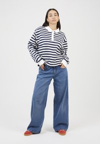 Woman standing with hands in pockets, wearing a navy and white striped long-sleeve polo, wide-leg jeans, and red sneakers.