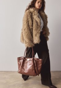 Manteau en fausse fourrure marron, haut blanc, pantalon noir. Porte un sac fourre-tout en cuir marron embossé crocodile avec une forme structurée et des doubles anses.