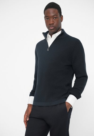 Sötét tengerészkék pulóver fél cipzárral és texturált anyaggal. A nadárban fehér ingujjak láthatók, sötét nadrággal párosítva.