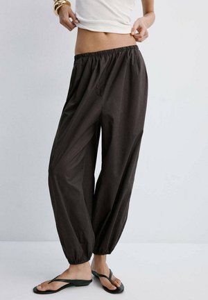 Mujer con pantalones negros holgados con cintura elástica, top blanco corto, pulseras doradas y chanclas negras de pie frente a un fondo liso.