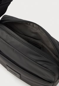 Fekete nylon keresztpántos táska cipzáros tetejével, bélés nélküli belső résszel és egy kis külső logóval. Simább felület és téglalap alak.