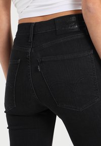 Jeans de denim negro con un ajuste slim, que presentan bolsillos traseros curvados y un parche de cuero en la cintura. Textura vertical sutil en la tela.