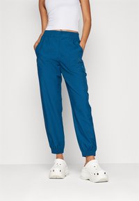 Modré volné kalhoty s elastickým pasem a manžetami, se dvěma předními kapsami. Nosené s bílými dřeváky.