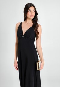 Schwarzes Abendkleid mit einem tiefen V-Ausschnitt und gerafftem Oberteil, das mit einer dekorativen Brosche versehen ist. Kombiniert mit einer rechteckigen goldenen Clutch.