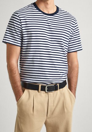 Mann trägt ein marineblau-weiß gestreiftes kurzärmliges Hemd, das in beige plissierte Hosen mit schwarzem Gürtel gesteckt ist, Hände in den Taschen, auf einfarbigem Hintergrund.