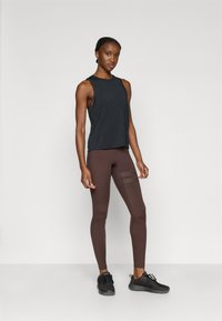 Μαύρο αμάνικο τοπ σε συνδυασμό με καφέ κολάν που διαθέτουν λεπτομέρειες από διαφανές δίχτυ, φορεμένα με μαύρα αθλητικά παπούτσια. Λεία υφάσματα, εφαρμοστό σχέδιο.