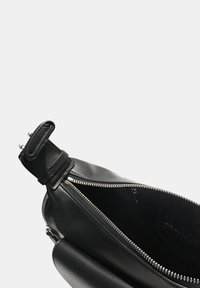 Sac en cuir noir avec fermeture éclair, présentant une texture lisse et un design épuré. Une sangle avec des éléments en métal est visible en haut.