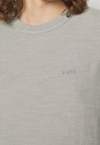 Nära bild av en person som bär en ljusgrå stickad tröja med ribbad rundhals och "BOSS" broderat på bröstet.