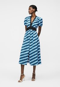 Γυναίκα με μπλε και μαύρο φόρεμα με μοτίβα, μήκους μέχρι τη μέση της γάμπας, με φουσκωτά μανίκια και ντεκολτέ σε σχήμα V, φοράει λευκά πέδιλα με ψηλό τακούνι, στέκεται με τα χέρια πίσω.