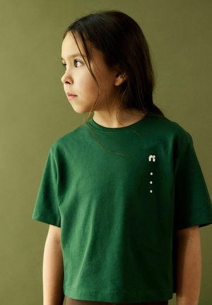 Young child with long dark hair wearing a green T-shirt with a small white logo, looking to the left against a muted green background.